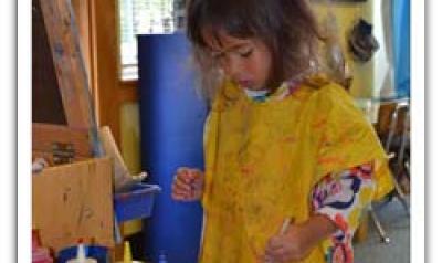 Child with paint smock on, standing near easel and paint containers.